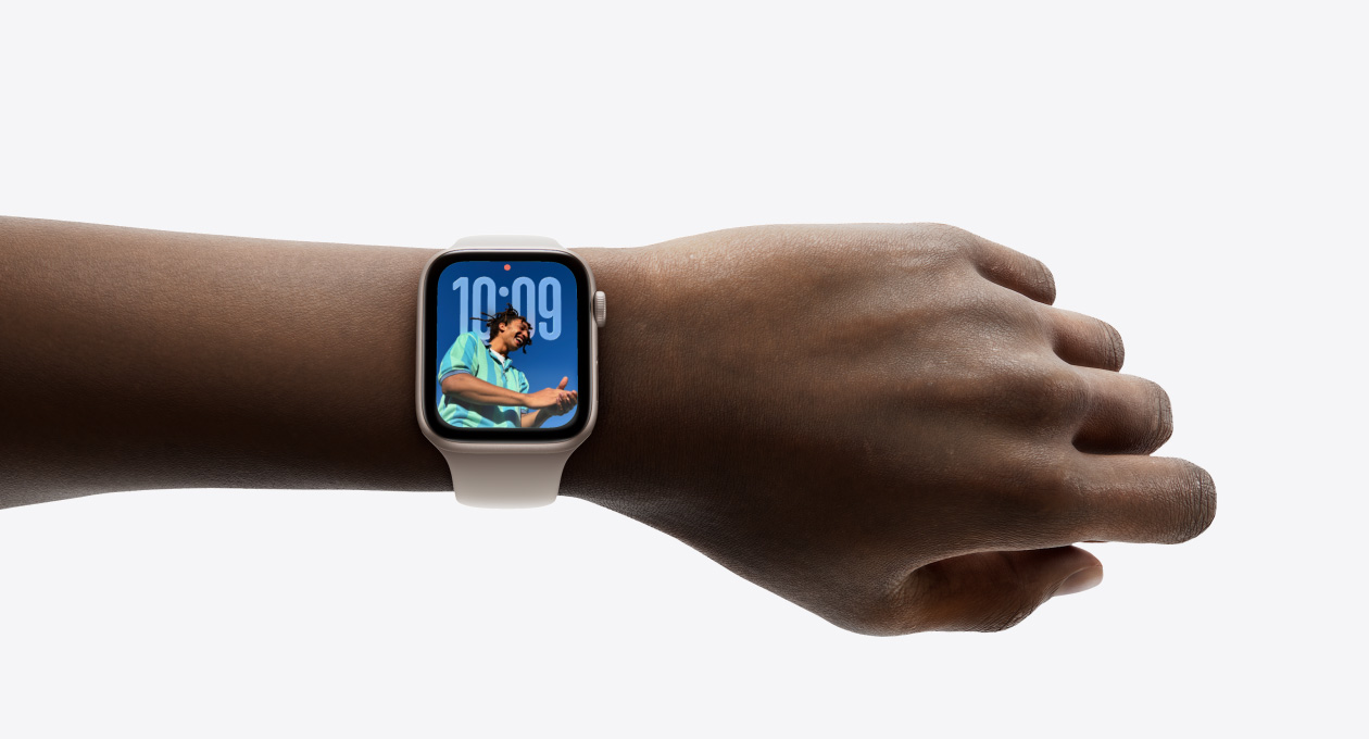 Close do Apple Watch com caixa prateada e pulseira de borracha no pulso de uma pessoa, mostrando o app Hora no novo design com Liquid Glass.