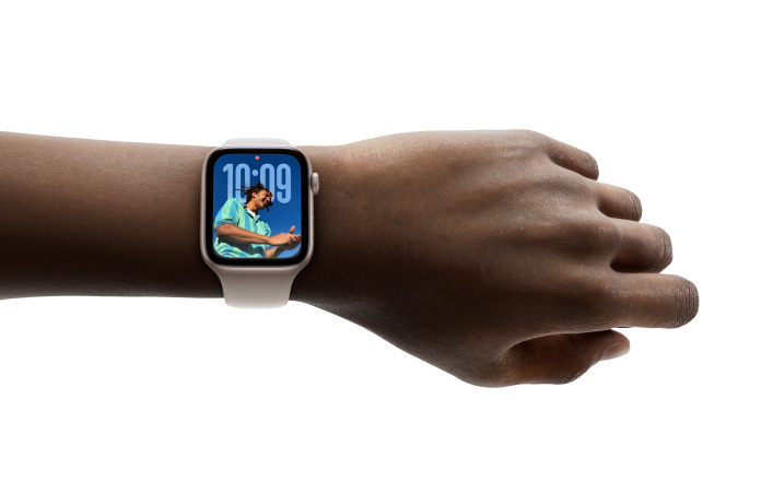 Gros plan d’une Apple Watch or rose avec un bracelet en caoutchouc au poignet d’une personne, qui affiche l’heure avec le nouveau design Liquid Glass