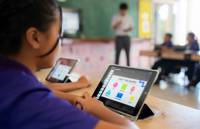 特寫一名學生坐在書桌前使用 iPad。螢幕顯示不同的彩色圖形，標題為「Meet the shapes」。