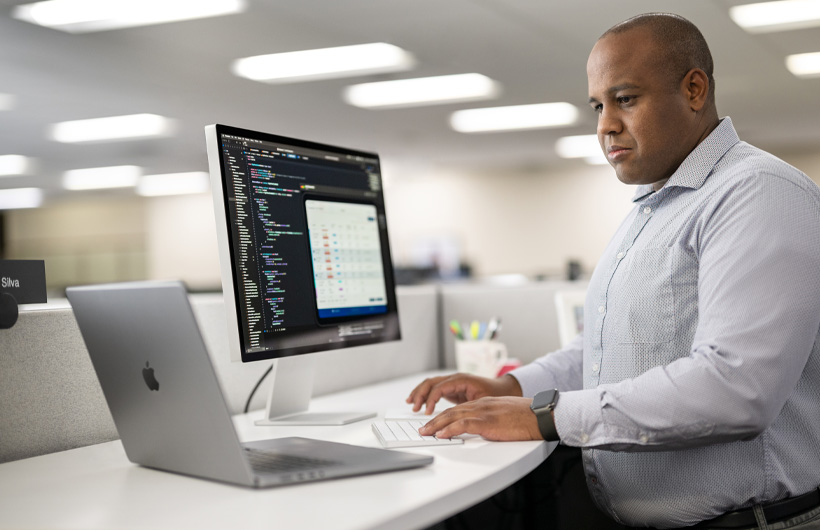 Un développeur debout à son bureau utilise un Mac et un MacBook; l’écran du Mac affiche du code et des informations relatives à une app