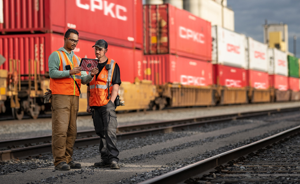 Deux cheminots consultent un iPad dans une gare de triage à côté d’un train immobilisé chargé de conteneurs
