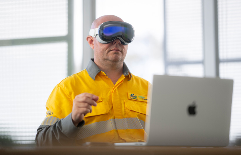 Un employé portant un Apple Vision Pro fait un geste de pincement; un MacBook Pro de couleur argent est posé devant lui, ouvert, sur le bureau
