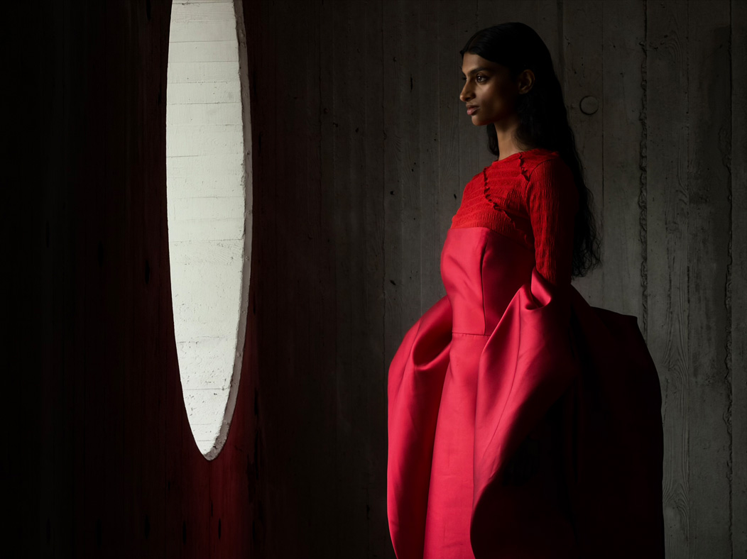 Eine Frau in einem langen roten Kleid steht in einem leeren Raum und wird fotografiert. Die Fotos App wechselt durch die Brennweiten, um die Möglichkeiten des Pro Kamera-Systems zu zeigen, hier: 200 mm