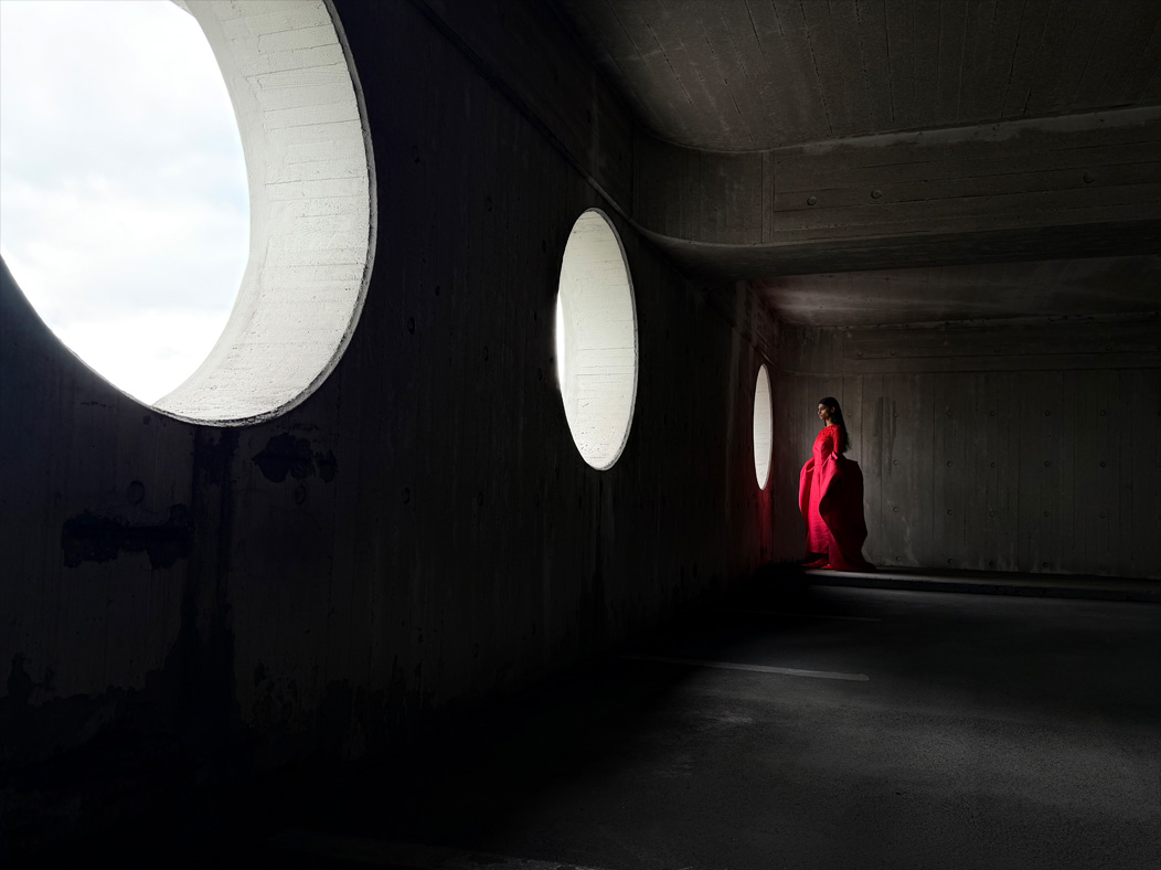 Eine Frau in einem langen roten Kleid steht in einem leeren Raum und wird fotografiert. Die Fotos App wechselt durch die Brennweiten, um die Möglichkeiten des Pro Kamera-Systems zu zeigen, hier: 28 mm